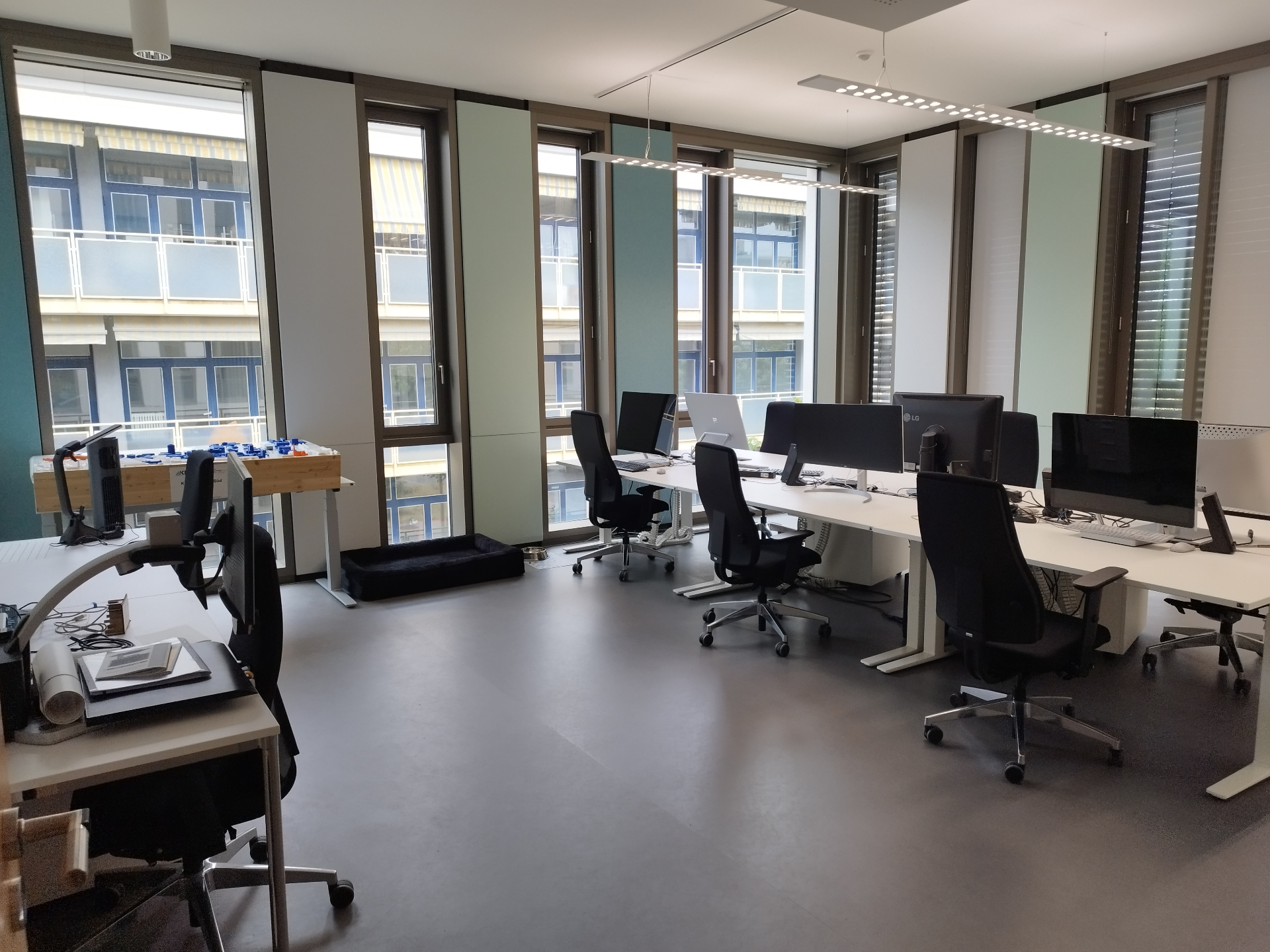 Der Studierendenraum des access@KIT im 1. Obergeschoss des InformatiKOM II. Ein heller, großzügiger Raum mit boden­tiefen Fenstern entlang der gesamten linken und hinteren Wand, die viel Tageslicht hereinlassen. In der Mitte stehen mehrere höhenverstellbare weiße Schreibtische mit ergonomischen schwarzen Bürostühlen, ausgestattet mit großen Bildschirmen und Surface-Studios. Im Vordergrund links ein Arbeitsplatz mit einem Bildschirmlesegerät und einem Ordner. An der hinteren Fensterfront steht ein Holztisch mit dem taktilen 3D-Campusplan. Auf dem grauen Boden liegt ein Hundekissen mit Napf.