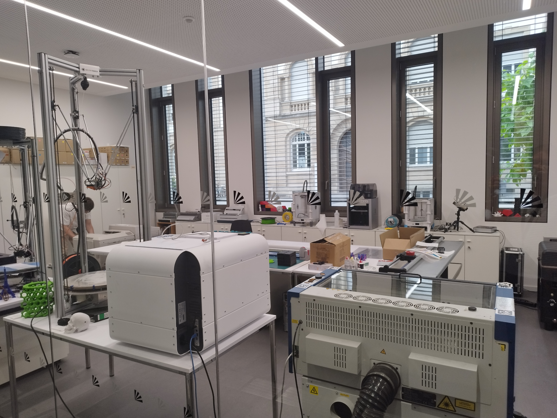 Das Drucklabor des access@KIT im Erdgeschoss des InformatiKOM II. Zu sehen sind mehrere 3D-Drucker, darunter ein großer Delta-Drucker mit Aluminiumrahmen auf der linken Seite, weitere Tischgeräte sowie ein Laserschneider im Vordergrund. An den Tischen entlang der hohen Fenster stehen verschiedene Filament-3D-Drucker und Materialproben. Im Hintergrund arbeitet eine Person an einem der Geräte.