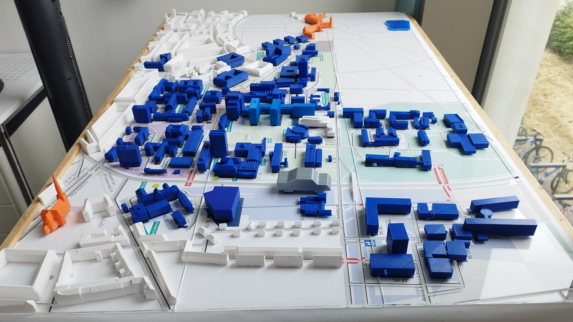 Der taktile 3D-Campusplan des KIT im Studierendenraum im 1. OG. Auf einem großen, schräg vor einem Fenster stehenden Tisch ist der gesamte KIT-Campus als ertastbares Modell aufgebaut. Die Gebäude sind als kleine, kräftig blaue 3D-gedruckte Blöcke dargestellt, einige besonders wichtige Gebäude in leuchtendem Orange. Straßen, Wege und Grünflächen sind als flache, beschriftete Grundfläche aufgedruckt – mit Straßennamen wie Adenauerring, Engler-Bunte-Ring und Hertz-Allee sowie Gebäudenummern. Im Hintergrund ist durch das Fenster eine begrünte Außenfläche zu erkennen.